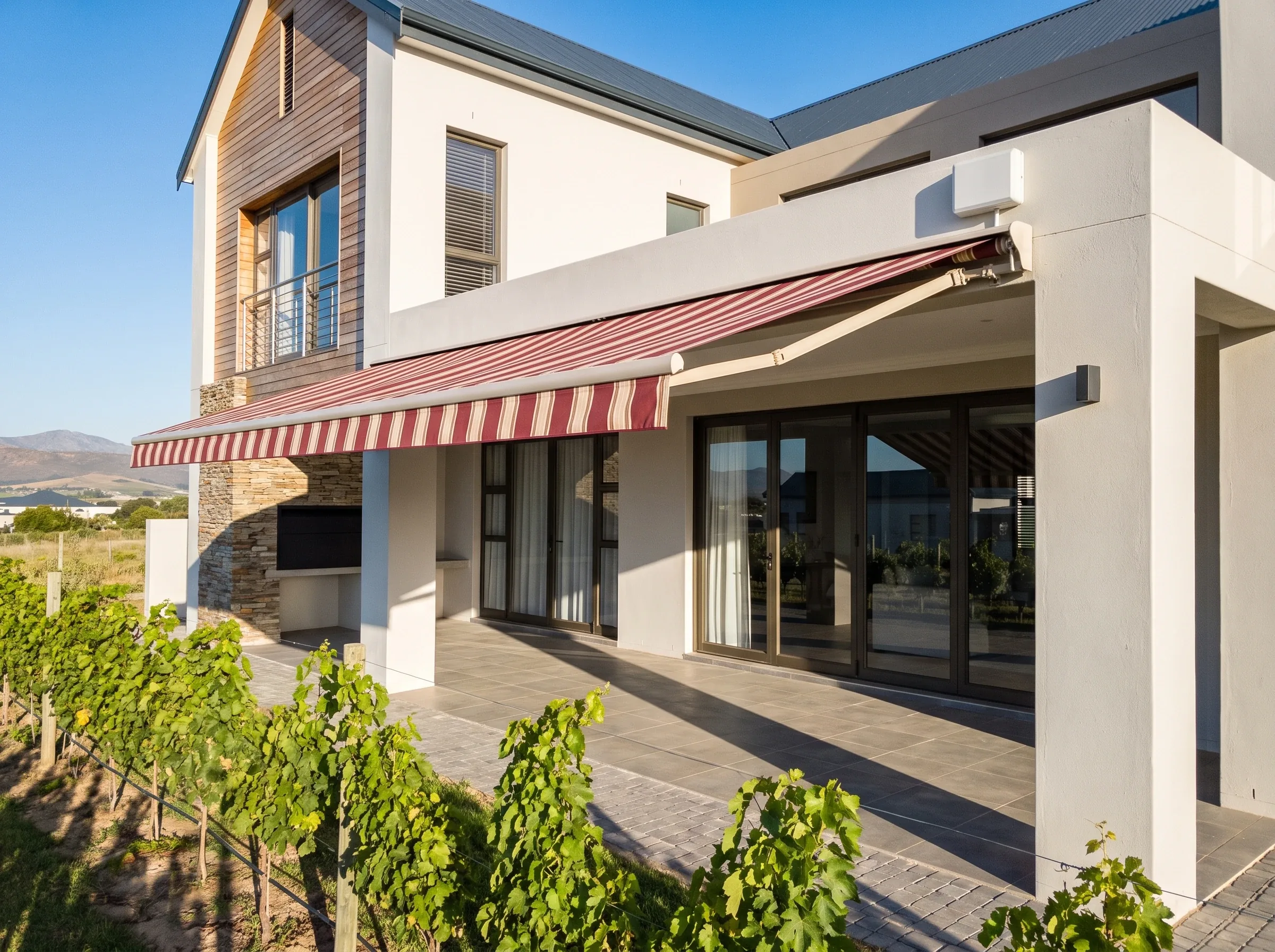 Contemporary double-storey Graanendal estate home with a striped folding-arm awning extended over a balcony, vineyard rows in the foreground
